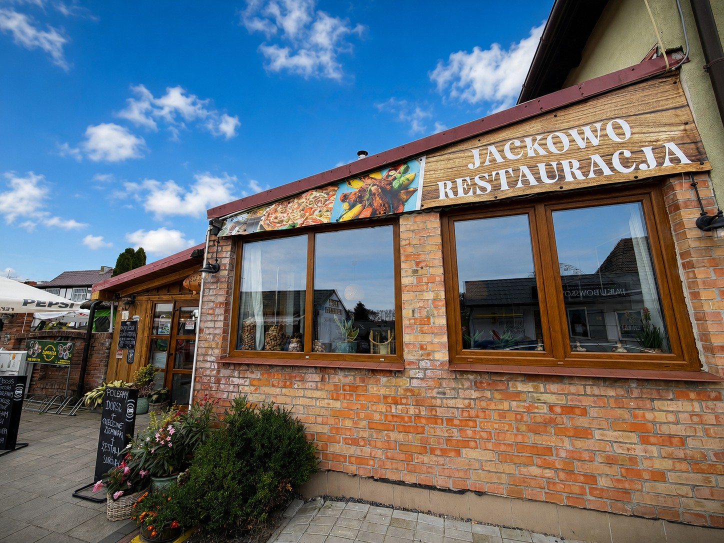 Front restauracji Jackowo w słońcu: drewniany szyld z napisem Jackowo Restauracja nad ceglaną ścianą, przed wejściem tablica z daniem dnia i kwiaty