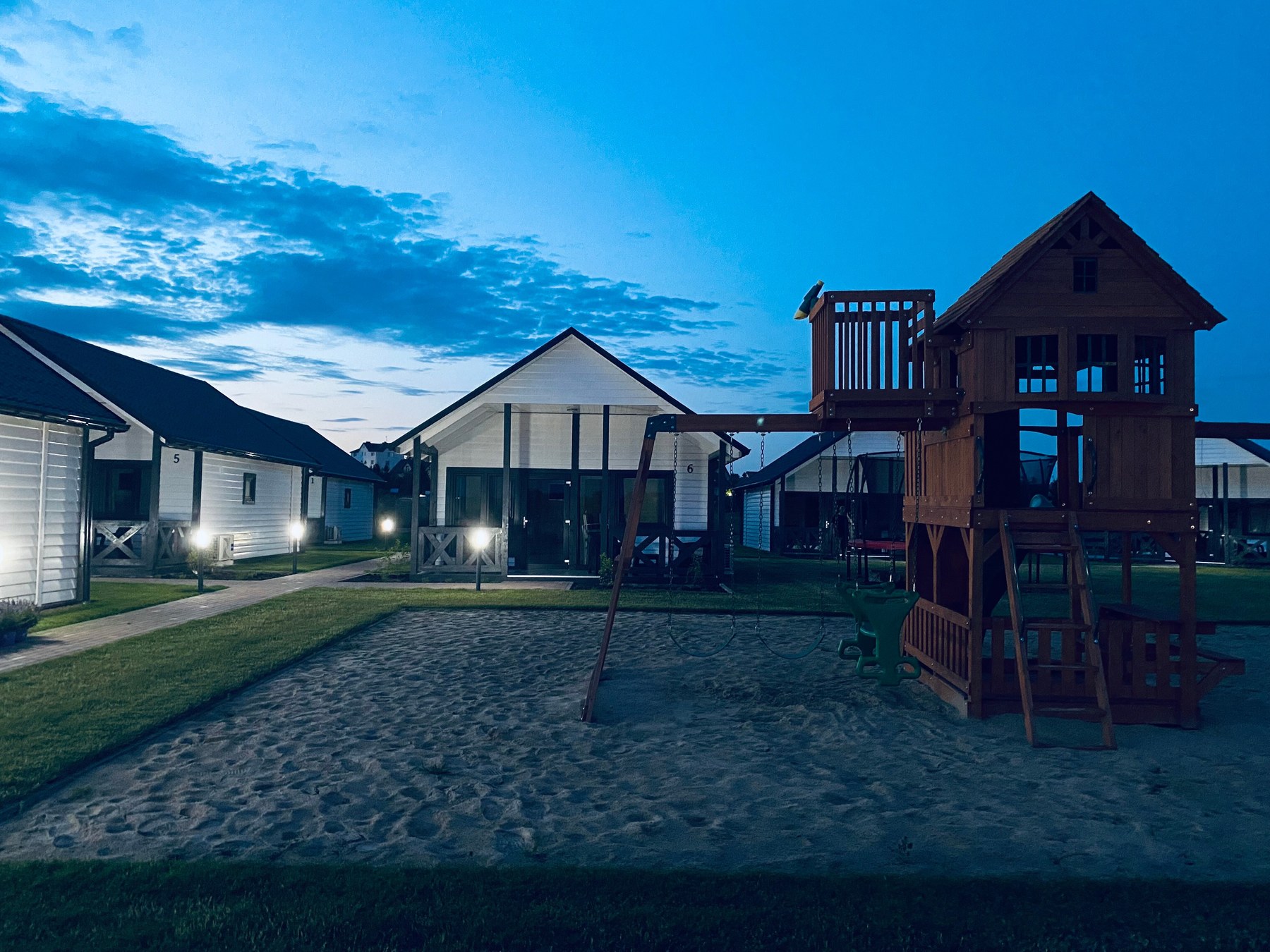 Plac zabaw z huśtawkami i drewnianą wieżą na piasku, w tle oświetlone domki o zmierzchu