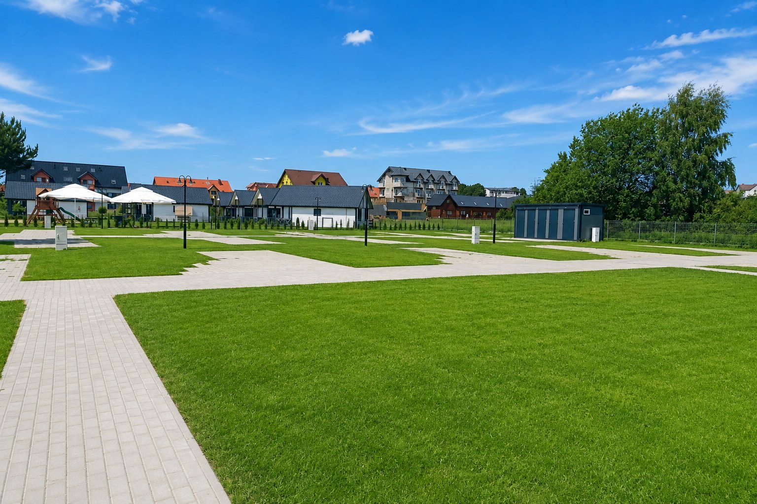 Pole kempingowe: utwardzone stanowiska w zielonej murawie, w tle domki osady
